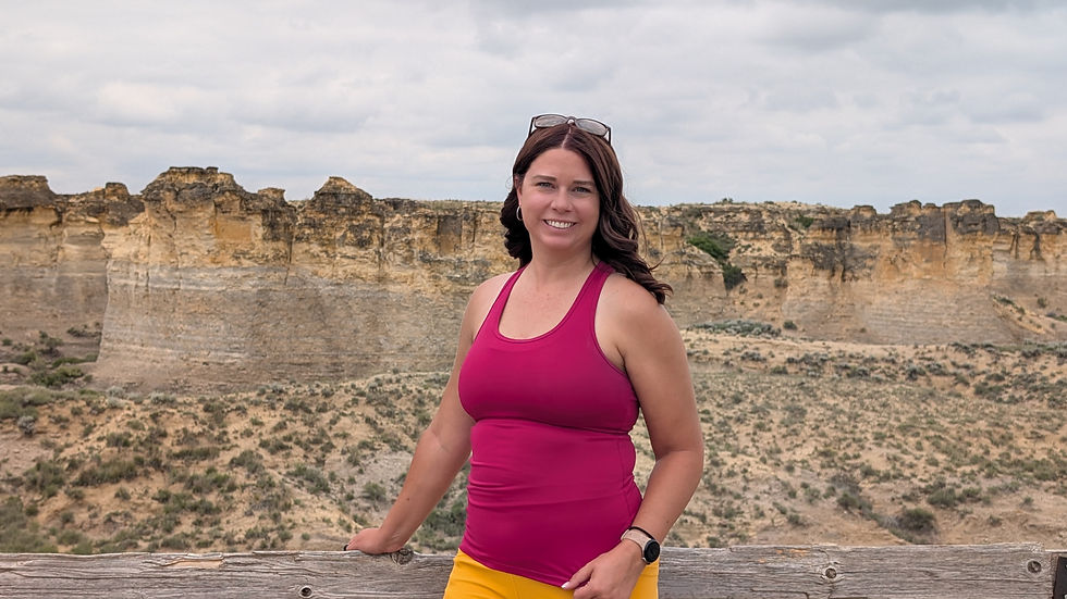 Smiling woman in a pink tank top stands at a wooden overlook railing above a rocky canyon under cloudy sky.