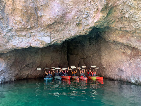 Kayakers raise paddles inside a rocky cave. They are in colorful kayaks on calm, turquoise water, creating a sense of adventure.