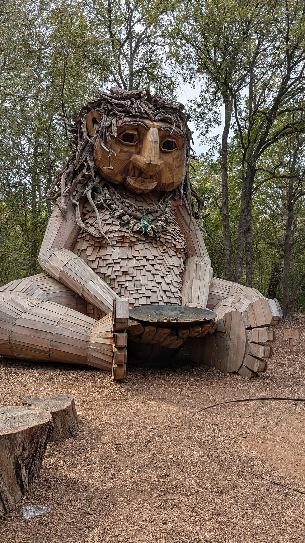 Wooden giant sculpture with leafy hair and stone necklace sits holding a bowl. Positioned in a forest clearing, surrounded by trees. Peaceful mood.