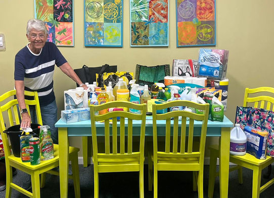 An older lady stands beside a table and chairs piled high with donated cleaning supplies and food.