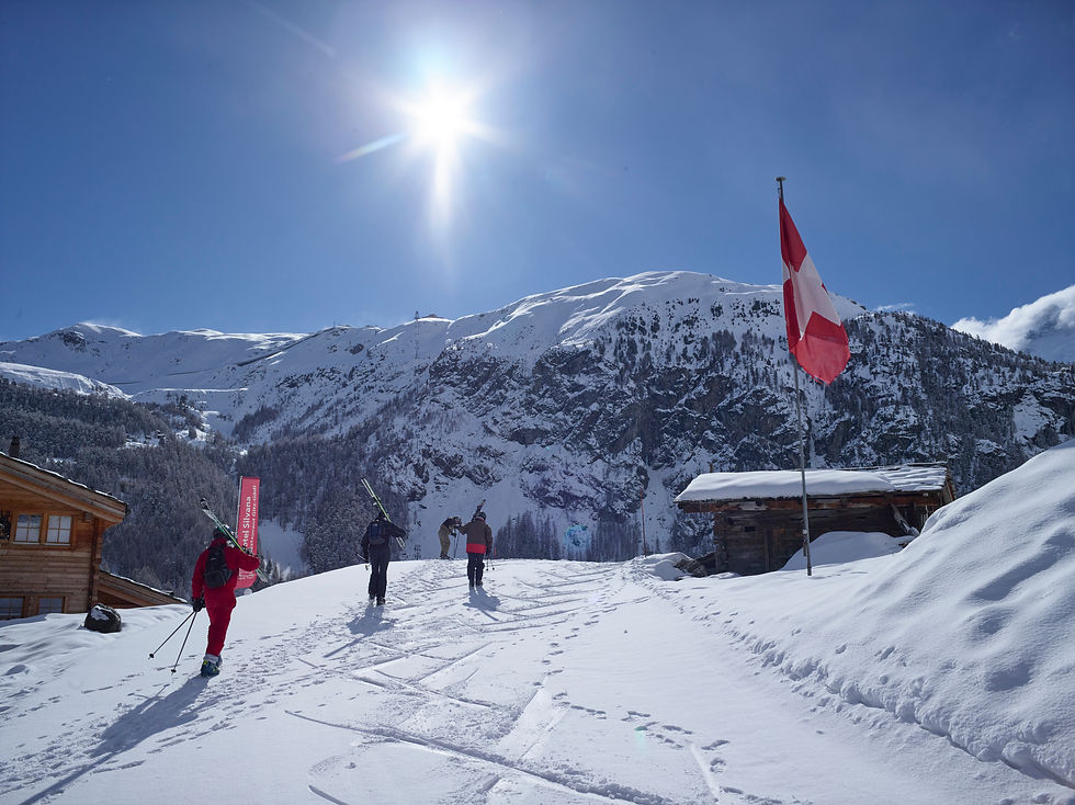 Landschaftsaufnahme eines sonnigen Skitags in den Schweizer Alpen mit Skifahrern im Vordergrund, einer Schweizer Flagge rechts und schneebedeckten Bergen im Hintergrund
