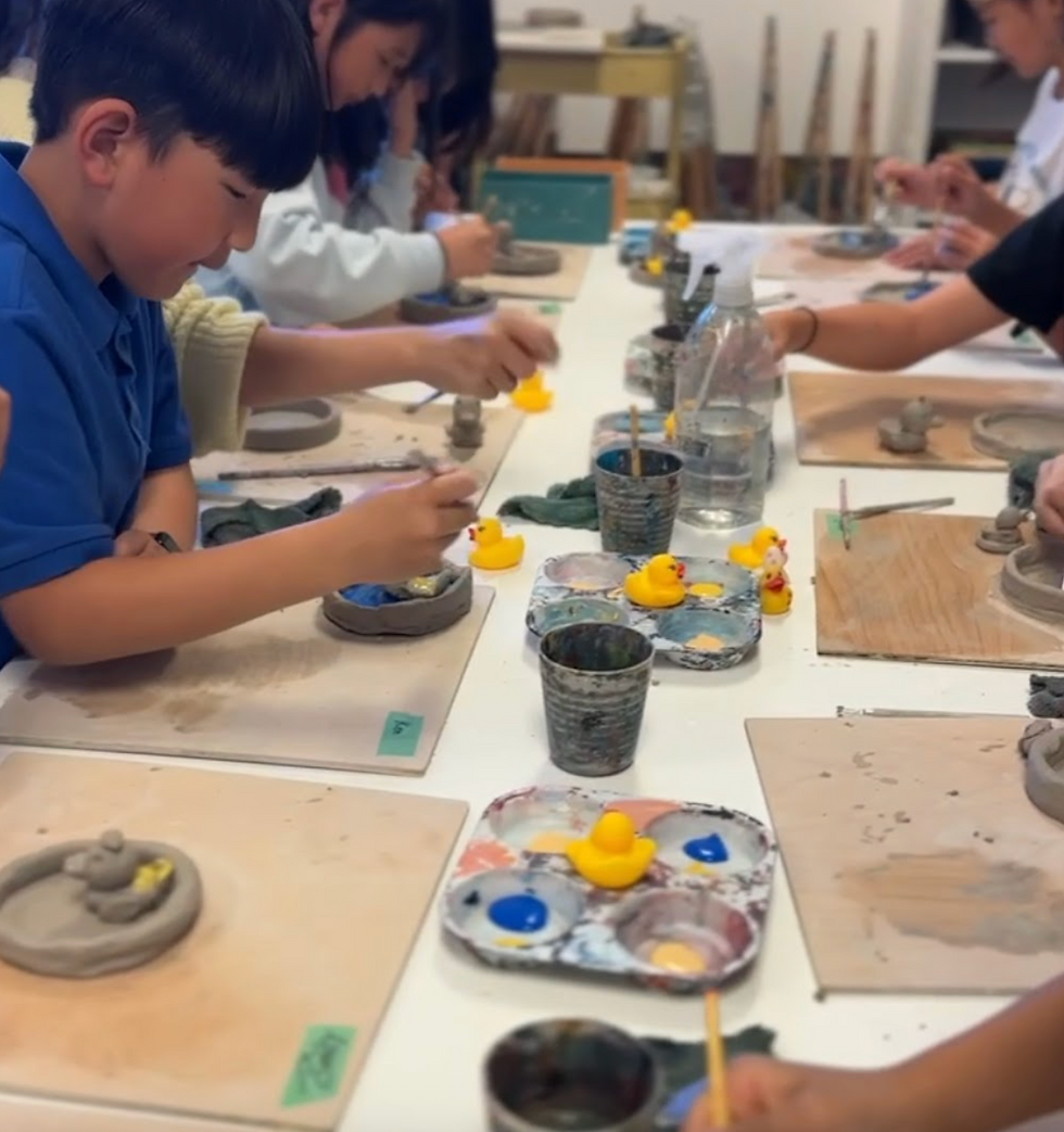 Children engaged in pottery painting at a table. Yellow rubber ducks and paint palettes visible. Focused and creative atmosphere.