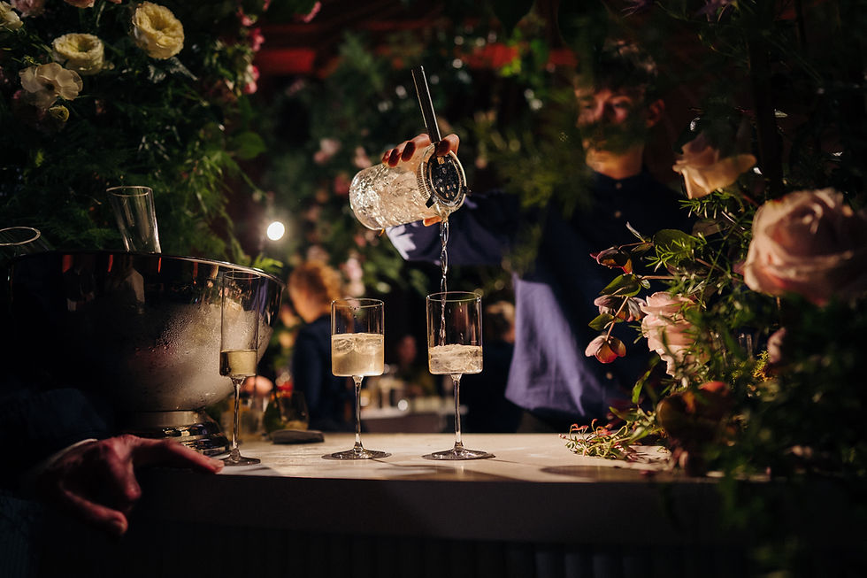 Bartender pours champagne into two flutes, behind a bar, WEDDINGS, floral background.