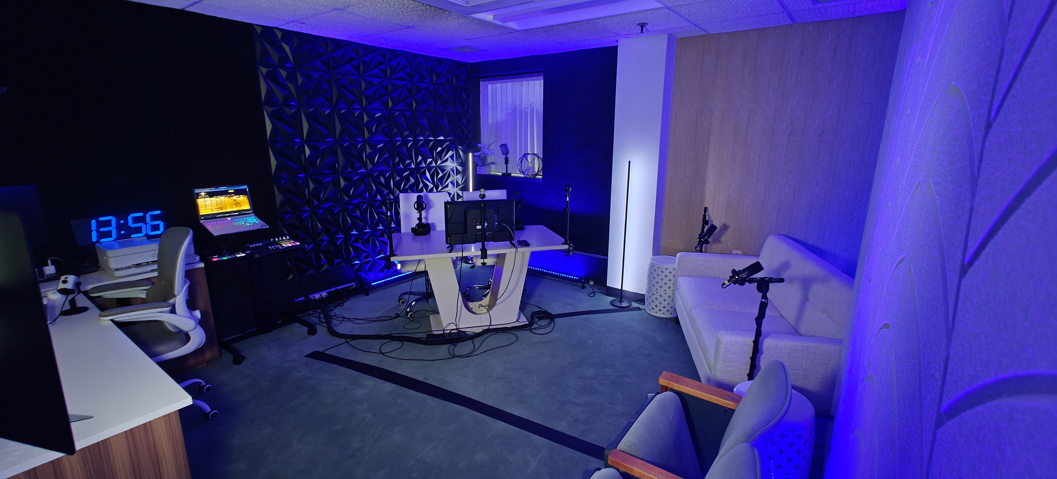 Wide view of the studio with blue accent lighting: producer desk with digital clock and monitors, white anchor desk in front of a geometric 3D wall panel, and gray armchairs.