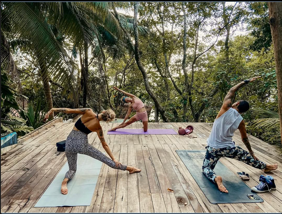 Tres personas haciendo yoga en una plataforma de madera en la selva. Vestimenta colorida, rodeados de árboles, ambiente sereno.
