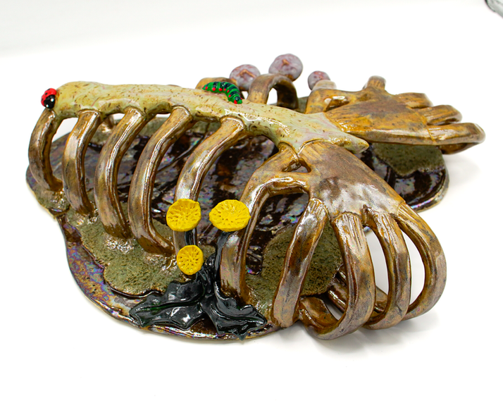 Contemporary ceramic sculpture “Cage of Bones” by Canadian artist Paige S. Harris. A large, golden-glazed rib cage emerges from a dark, rainbow-glistening puddle, with moss growing at the base of the bones. Dandelions and mushrooms, along with a ladybug and a caterpillar, make their home on top.