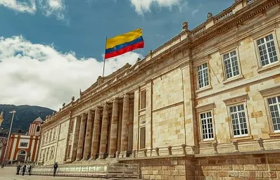 Bandera de Colombia ondeando sobre un edificio neoclásico de piedra clara, con columnas y cielo nublado al fondo.