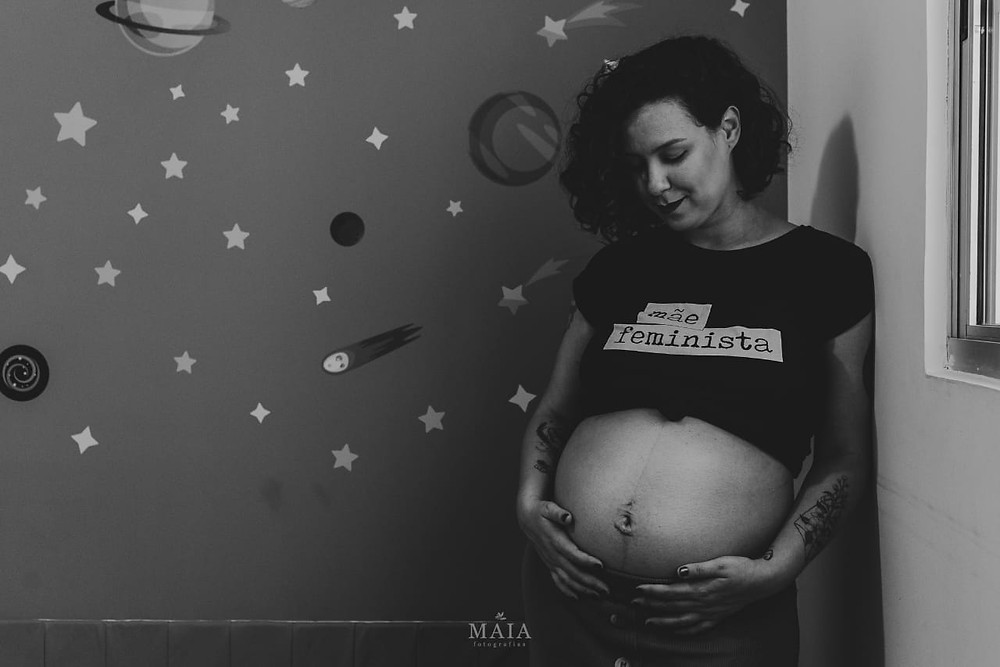 Cris usa camiseta preta escrito mãe feminista e acaricia a barriga de grávida. Ela olha para baixo e sorri. Atrás dela, uma parede de estrelas e planetas
