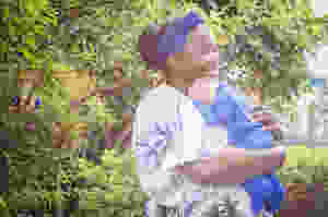 Na frente de um jardim com árvores e arbustos, Liliane segura Benício no colo. O bebê dorme tranquilo, e ela sorri repousando suavemente o rosto sobre a cabecinha do bebê