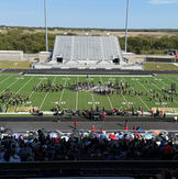 Marching band performs on sunny football field before large stadium audience.