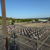 Students practice marching band formation on a large paved lot.