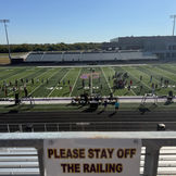 Marching band practices on football field beneath "PLEASE STAY OFF THE RAILING" sign.