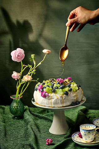 A styled food photograph of a pavlova topped with blueberries, green grapes, and pink edible flowers on a white cake stand, with a hand drizzling honey from a gold spoon. Pink ranunculus flowers in a green glass vase and a blue and white teacup are arranged alongside on a draped green linen cloth.