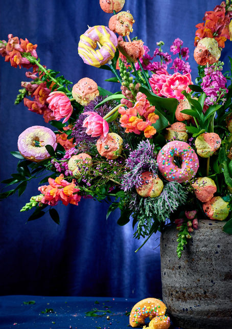 A vibrant floral arrangement of tulips, snapdragons, and wildflowers interwoven with colorful glazed donuts and cake pops on stems, displayed in a speckled stone vase against a deep cobalt blue background — bold editorial food photography by Peggy Cormary.