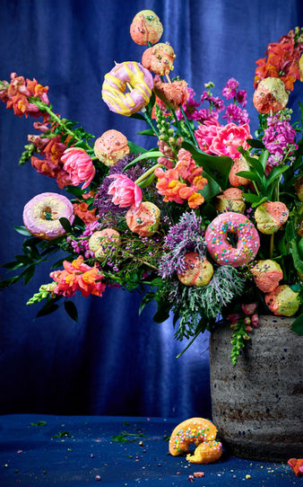 A vibrant floral arrangement of tulips, snapdragons, and wildflowers interwoven with colorful glazed donuts and cake pops on stems, displayed in a speckled stone vase against a deep cobalt blue background — bold editorial food photography by Peggy Cormary.