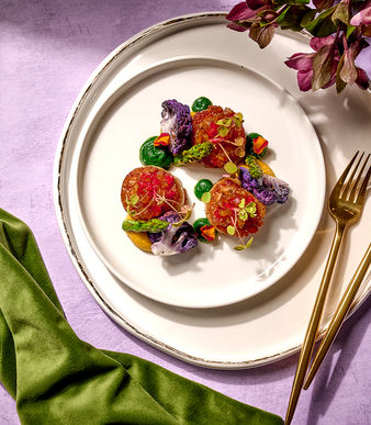 Overhead view of a refined seasonal dish with colorful vegetables and garnishes on white ceramic plates, styled with gold flatware and florals on a lavender backdrop