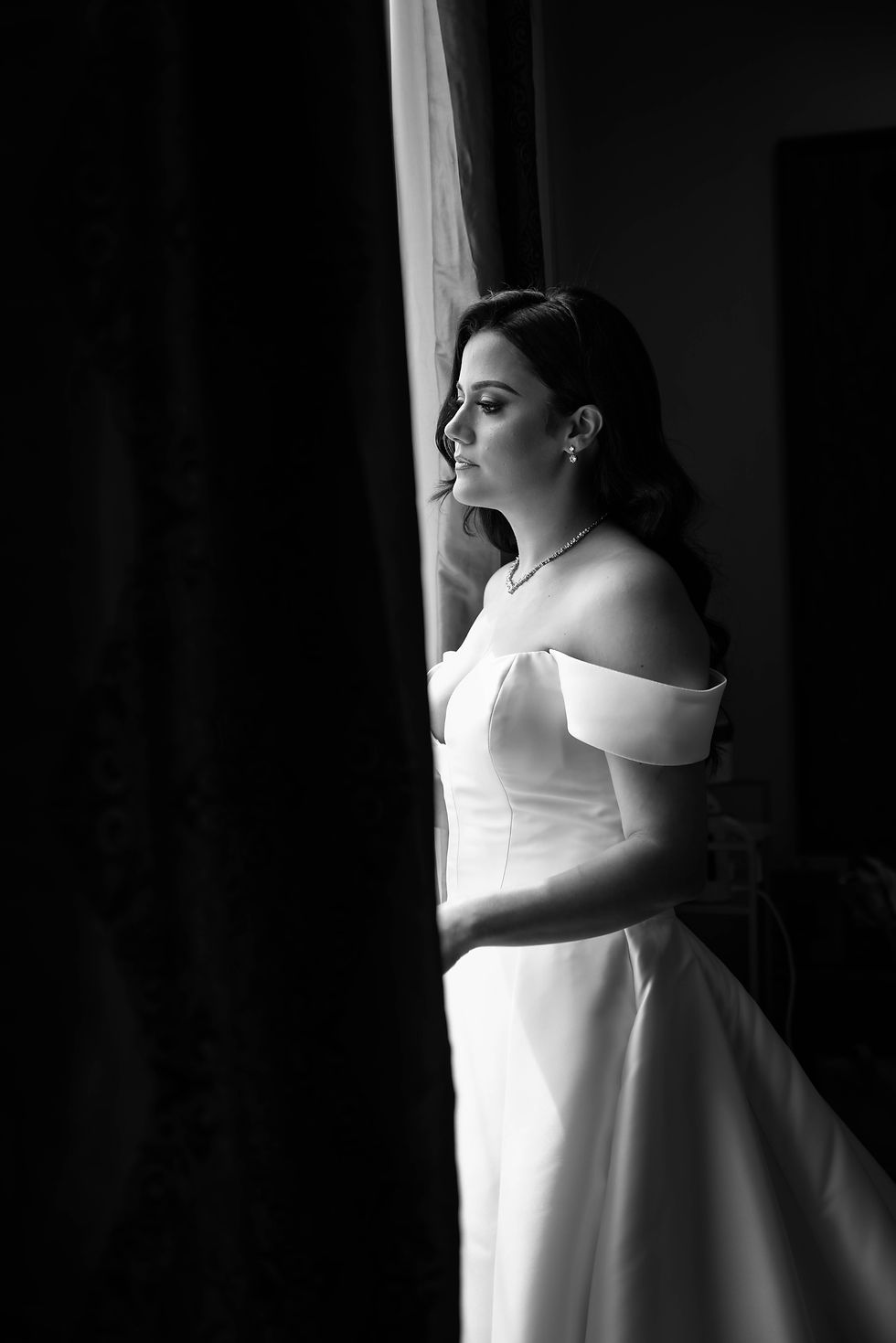 Bride gazes out a window in a dim room, wearing an off-shoulder white gown. Light highlights her serene expression and wavy hair.