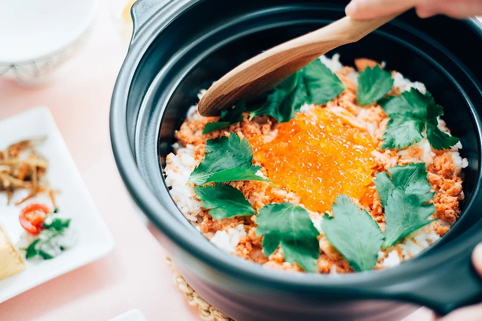 Hand stirring rice topped with salmon, roe, and green leaves in a black pot. Side dish with veggies on the left. Bright, appetizing scene.