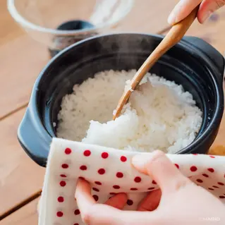 Hands serving steaming rice from a Hario Rice Cooker with a wooden spoon, using a white cloth with red dots. Warm, cozy kitchen setting.
