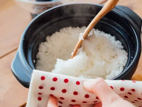 Hands serving steaming rice from a Hario Rice Cooker with a wooden spoon, using a white cloth with red dots. Warm, cozy kitchen setting.