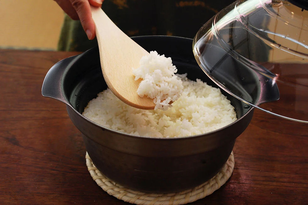 Hand scooping steamed rice with a wooden spatula from a black pot on a wooden table. The Hario Induction Rice Cooker has a glass lid, and the mood is warm.