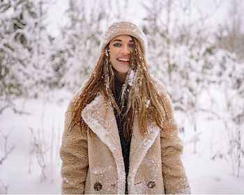 Junge Frau im Winter in verschneiter Natur, trägt eine warme Mütze und einen Wollmantel, während Schnee auf Haaren und Kleidung liegt. Das Bild symbolisiert die Belastung der Haare durch Kälte, trockene Winterluft, Mützen und äußere Umwelteinflüsse – typisch für die Wintermonate, in denen Haare Feuchtigkeit verlieren, matt und spröde werden und Schutz sowie intensive Pflege benötigen, bevor der Frühling beginnt. Jetzt Termin buchen bei Balayage Pro Studio in Konstanz