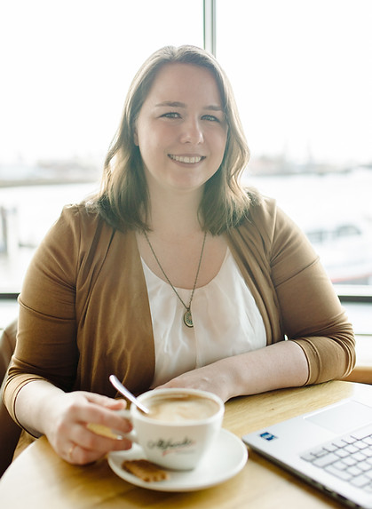 Annika sitzt mit einem Kaffee und Laptop an einem Holztisch. Im Hintergrund sind die Landungsbrücken zu erahnen.
