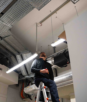 Monteur Alexaksander blickt an die Decke auf einen Rauchmelder, Seitenschneider in der Hand – Aufnahme während der Installation im Technikraum