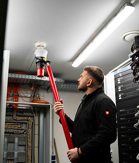Aleksander prüft mit einer Melderstange einen Rauchmelder an der Decke – Wartungsvorgang im Technikraum bei JK Building Protection