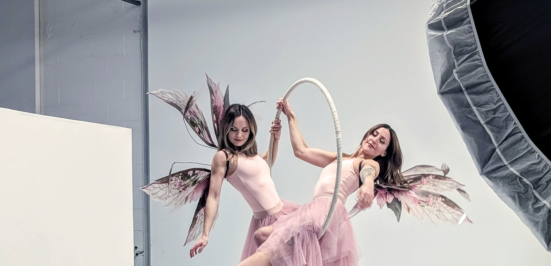 Two women dressed as fairies with pink wings and tulle skirts posing on an aerial hoop in a white cyclorama studio, with floral decorations around the hoop base and studio lighting equipment visible on the side.