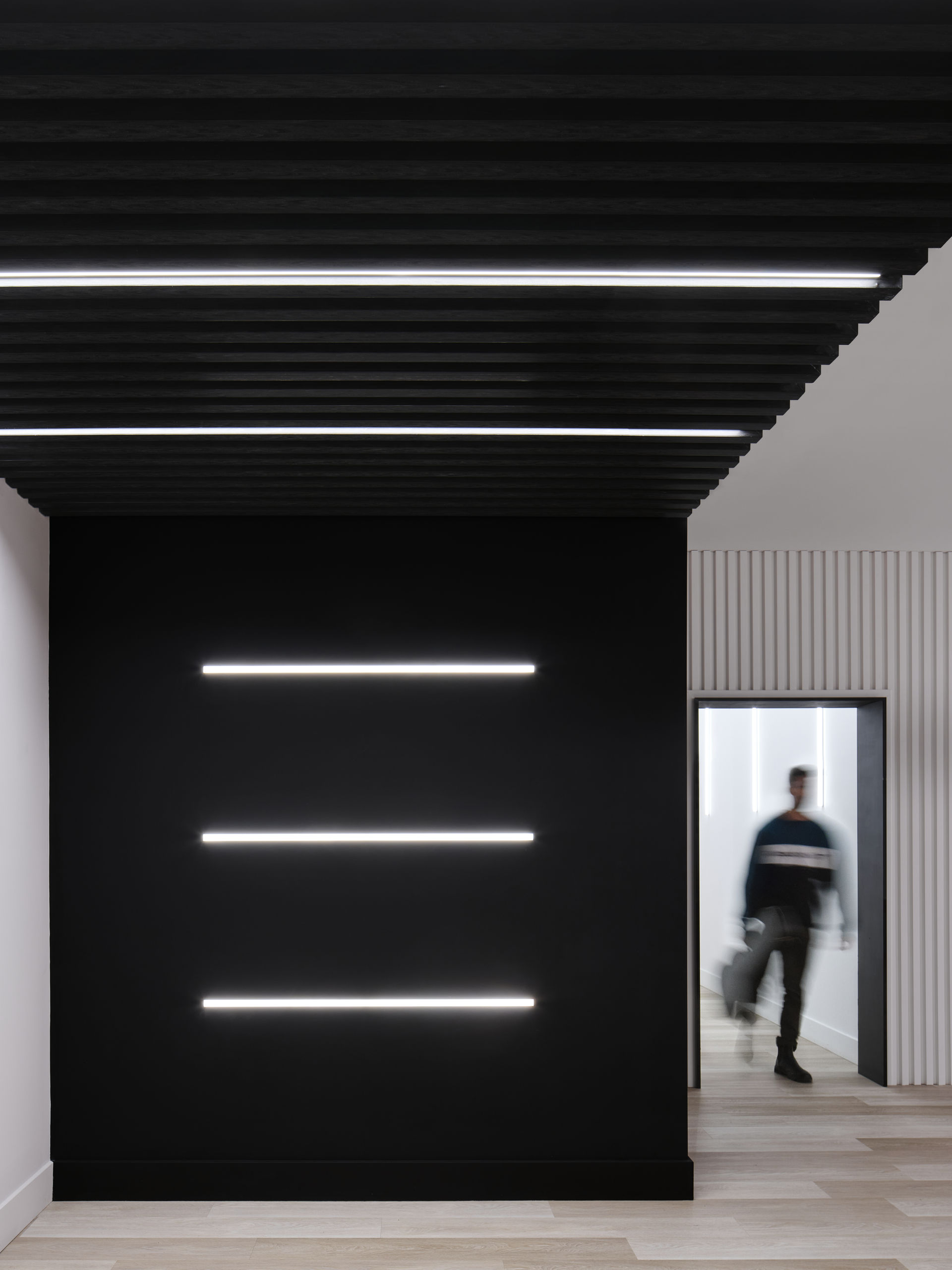 Modern hallway with black acoustic slat ceiling and horizontal LED light strips, matte black feature wall, and a person walking through the doorway in soft motion blur.