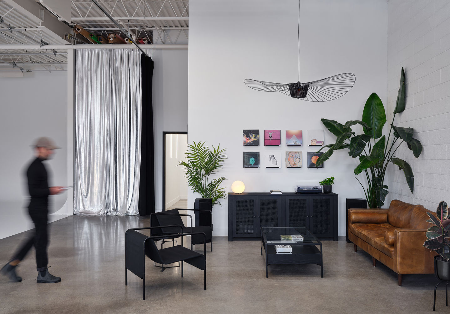 Studio lounge with a leather sofa, black chairs, artwork display, greenery, and a silver curtain leading to the main shooting area.