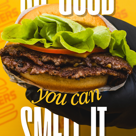 A gloved hand holds a juicy burger with lettuce on a yellow backdrop. Text reads “So good you can smell it.” Bold, appetizing mood.