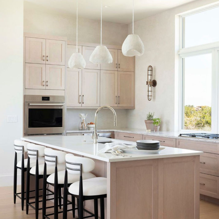 Kitchen with light wood cabinetry, a large white island, upholstered counter stools, sculptural pendant lights, and tall windows.