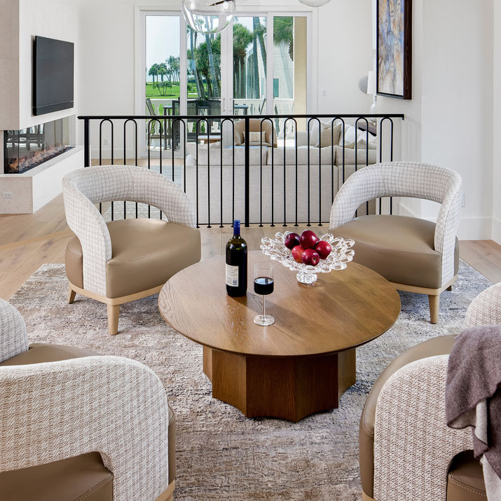 Loft seating area with curved upholstered chairs, round wood table, black railing, linear fireplace, and balcony doors with palm views.