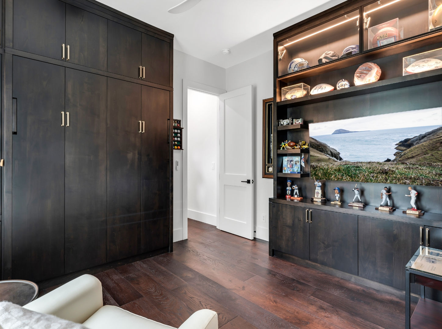 Home office or media room featuring floor-to-ceiling dark wood built-ins with illuminated sports memorabilia displays, integrated storage cabinetry, a wall-mounted television, and hardwood flooring.