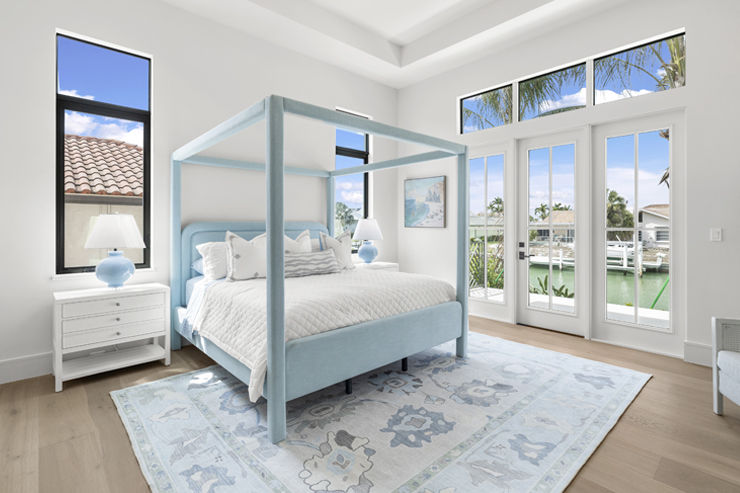 Bedroom with pale blue canopy bed, white bedding, matching blue lamps, patterned area rug, tall windows, and glass doors overlooking the water.