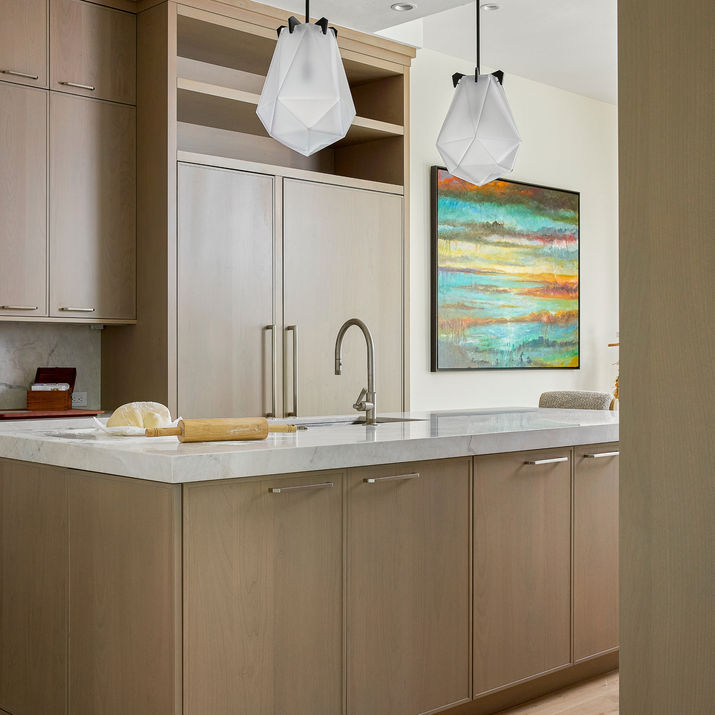 Kitchen detail with light wood cabinetry, marble countertop, island sink, geometric pendant lights, colorful artwork, and dough with a rolling pin.