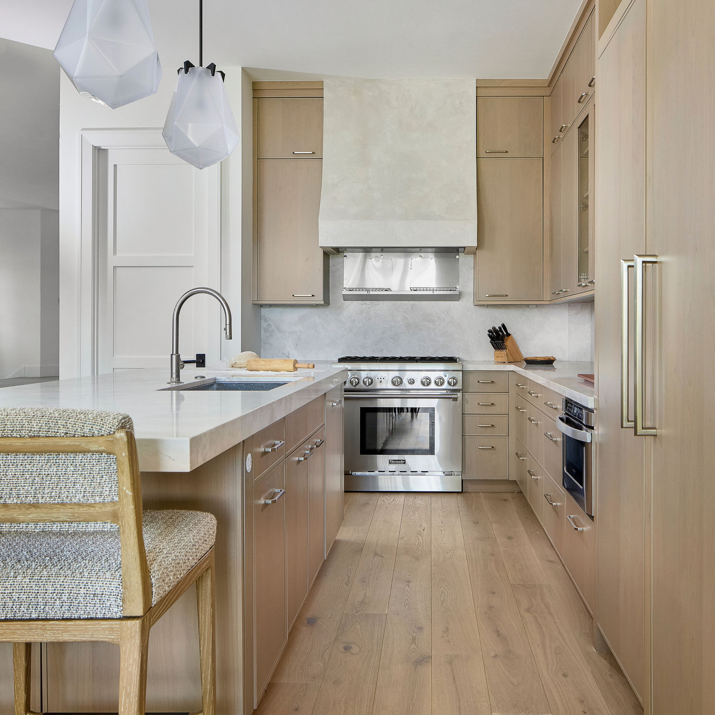 Kitchen with light wood cabinetry, marble countertops, stainless steel range, custom range hood, island sink, geometric pendant lights, and pale wood flooring.