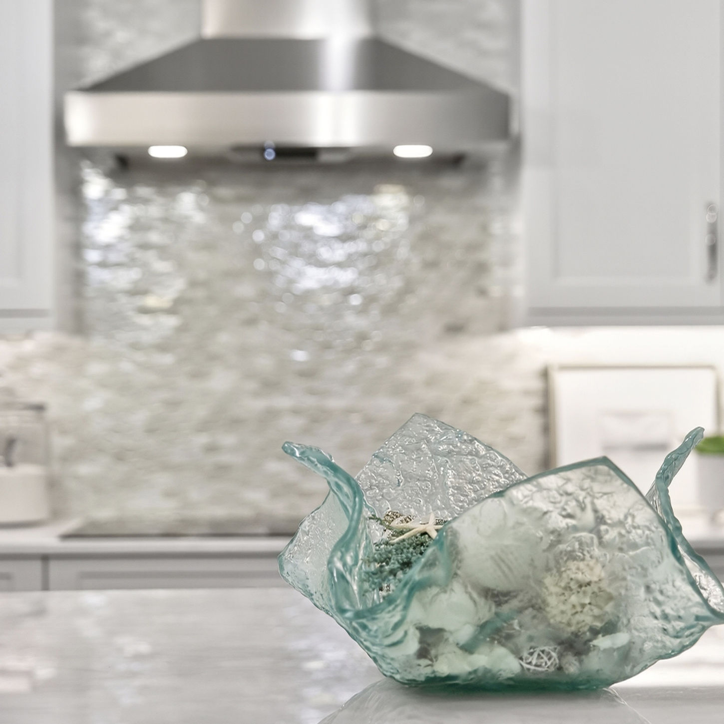 Kitchen detail featuring a sculptural aqua glass bowl filled with shells and coral, with a textured backsplash and stainless steel range hood beyond.
