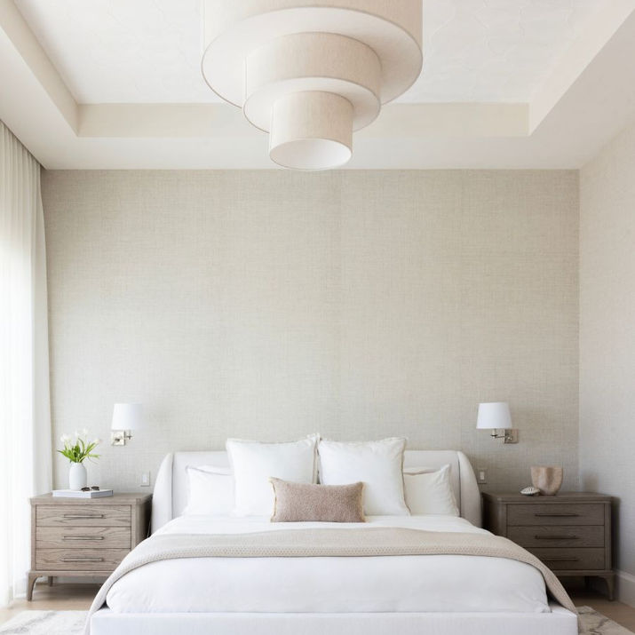 Primary bedroom with an upholstered white bed, layered neutral bedding, wood nightstands, textured wallcovering, and a tiered fabric chandelier.