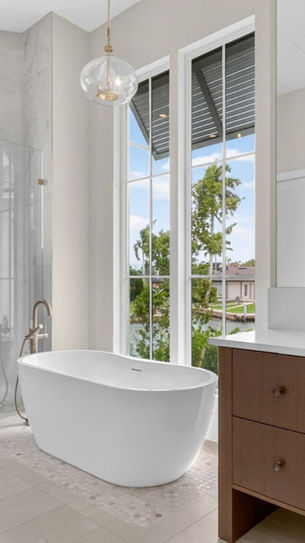 Modern luxury bathroom with freestanding tub, glass shower, wood vanity, and tall windows showing palm trees and water.