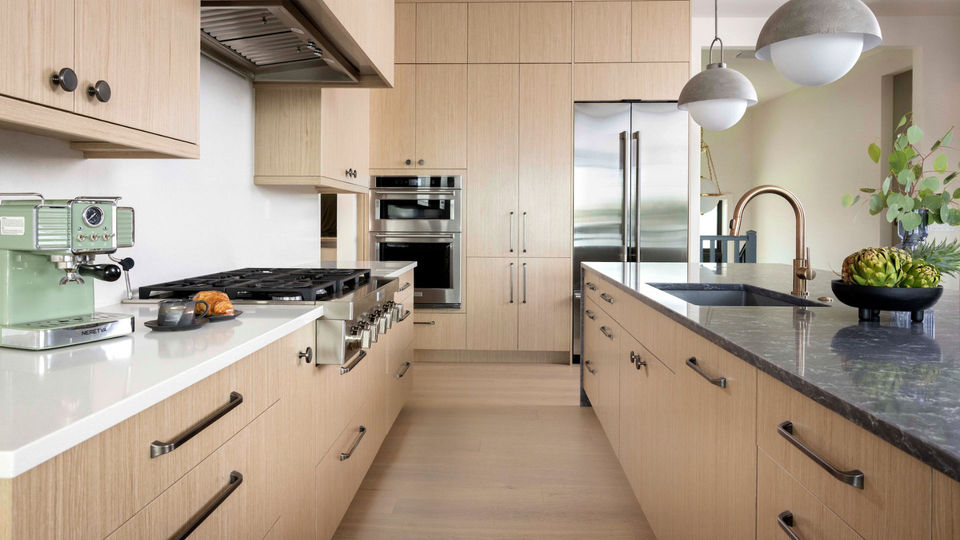 Modern kitchen with light wood cabinetry, professional appliances, a dark stone island, globe pendant lights, and a mint green espresso machine.