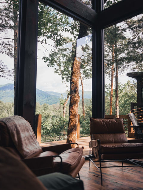 A brown leather chair sitting in front of tall, dark-framed windows looking out onto a bright green forest canopy and distant mountains. Architecture by James Knight Photography by Kalyn Kight. Reynard Architectural Designs