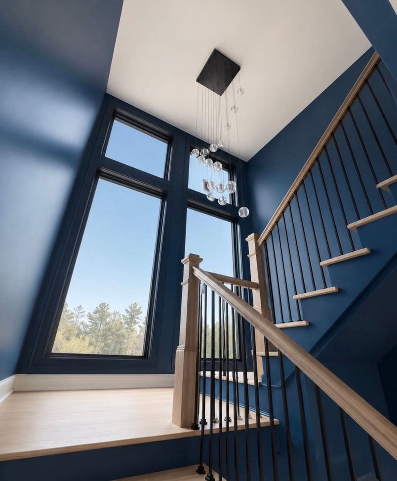 Modern stairwell with deep blue walls, tall windows, and a hanging glass chandelier in a bright, airy interior. Architecture by James Knight (Reynard Architectural Designs) Photo by Blue Dog Construction
