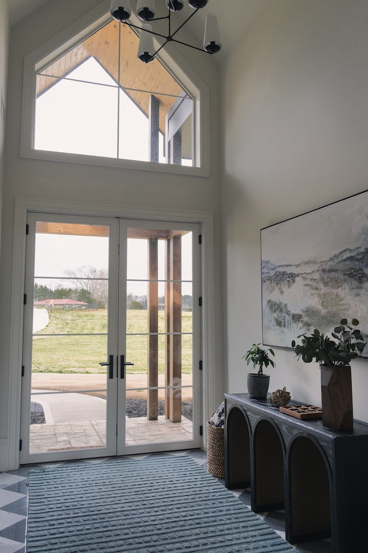 Tall modern entryway with double glass doors, arched window, blue rug, console table, plants, and landscape view outside. Reynard Architectural Designs Powder Springs, GA