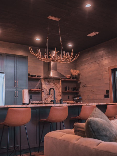 A modern rustic kitchen with gray cabinets, stone countertops, and bar seating, opening up to a living room with a large cozy sofa and an antler chandelier. Architecture by James Knight Photography by Kalyn Kight. Reynard Architectural Designs