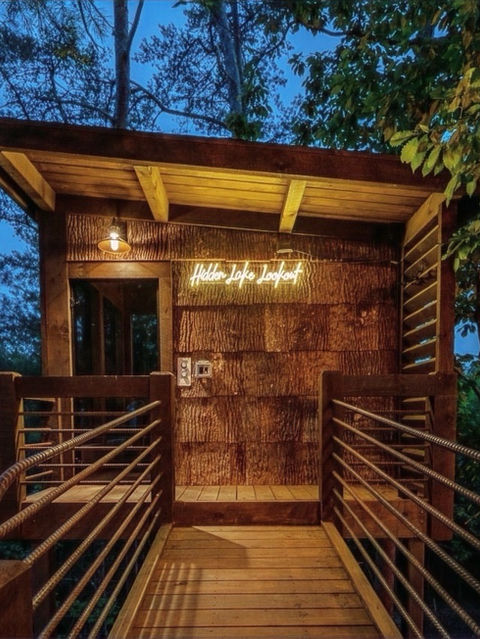 A wooden footbridge leading to a treehouse detached sauna entrance with textured bark siding, featuring a bright neon sign reading Hidden Lake Lookout against a twilight sky.Architecture by James Knight Photography by Kalyn Kight. Reynard Architectural Designs