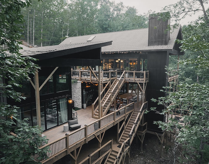 A striking, multi-story modern home nestled in a dense forest, featuring dark vertical siding and expansive, multi-level wooden decks connected by outdoor staircases. Large floor-to-ceiling glass windows reveal a warm, lit interior, while string lights hang above an upper outdoor terrace. A person walks along a middle walkway, illustrating the grand scale of the retreat against the surrounding trees.