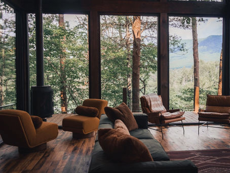 Cozy cabin lounge with brown chairs and sofa facing floor-to-ceiling windows overlooking a forested mountain view. Hidden Lake Lookout Cherry Log, GA Reynard Architectural Designs